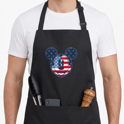 Funny americana fourth of july aprons - Mickey Mouse head silhouette filled with blue stars and red white stripes — shown on a black cotton canvas apron against a white background