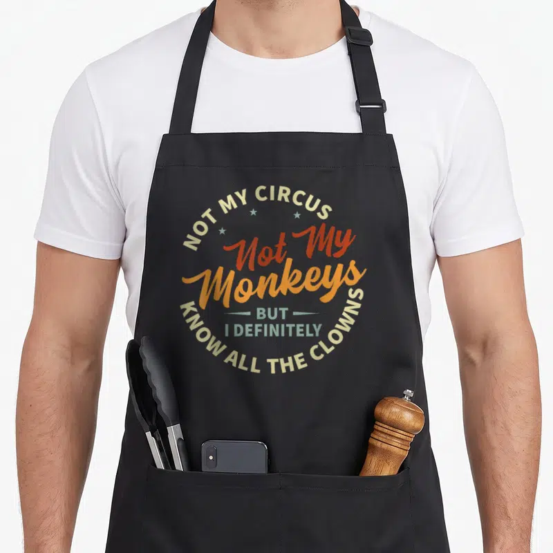 Irony weird aprons - Circular typographic design reading Not My Circus Not My Monkeys But I Definitely Know All — shown on a black cotton canvas apron against a white background