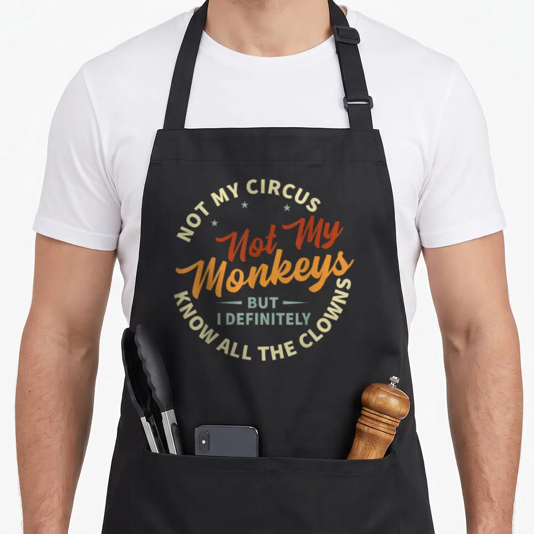 Irony weird aprons - Circular typographic design reading Not My Circus Not My Monkeys But I Definitely Know All — shown on a black cotton canvas apron against a white background