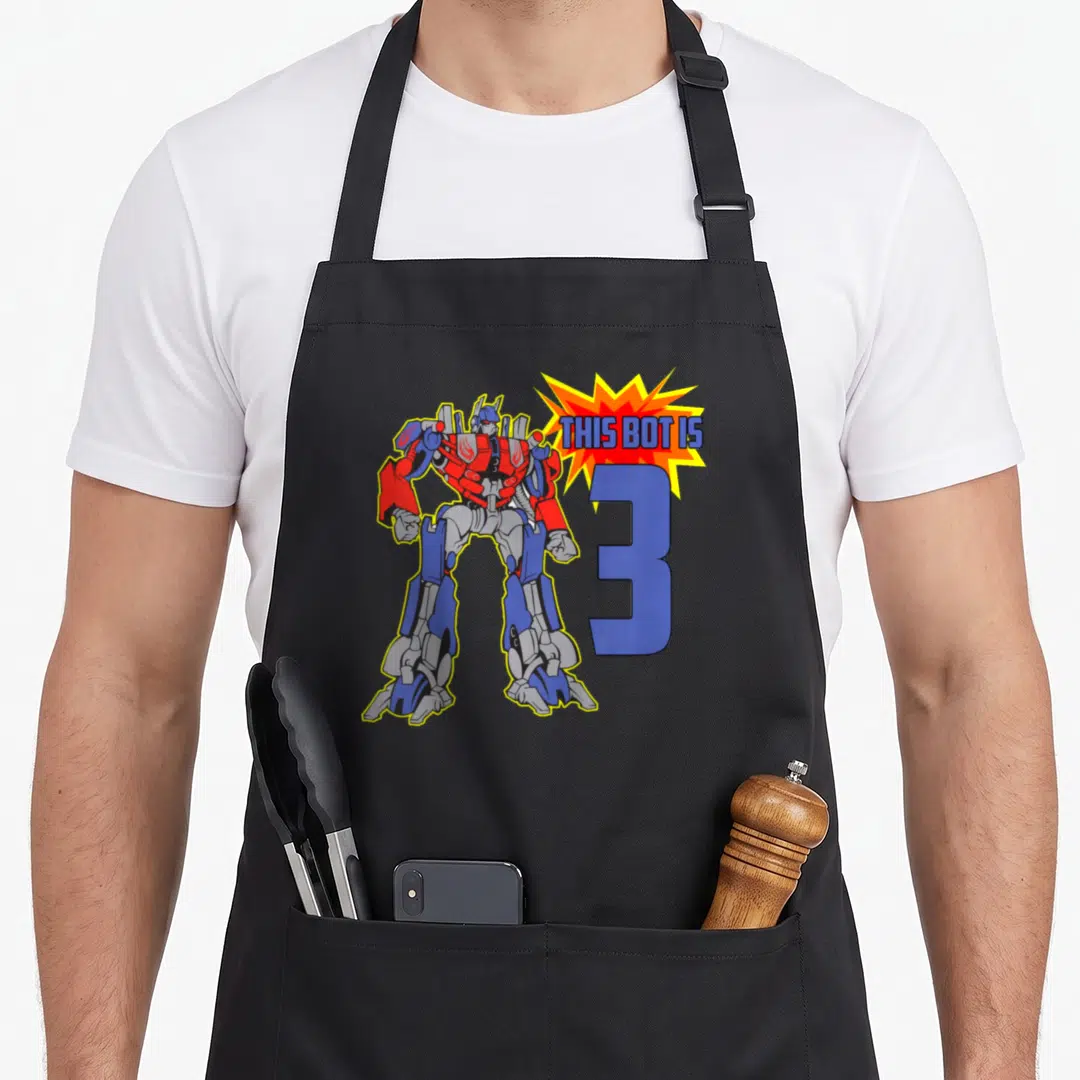 3rd bot birthday apron - Cartoon robot in red blue grey stands by blue number 3 under yellow text burst — shown on a black cotton canvas apron against a white background