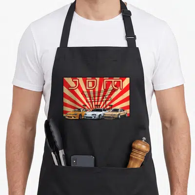 Jdm japanese car apron - Three cartoon cars under a red torii gate with JDM text on a sunburst background — shown on a black cotton canvas apron against a white background
