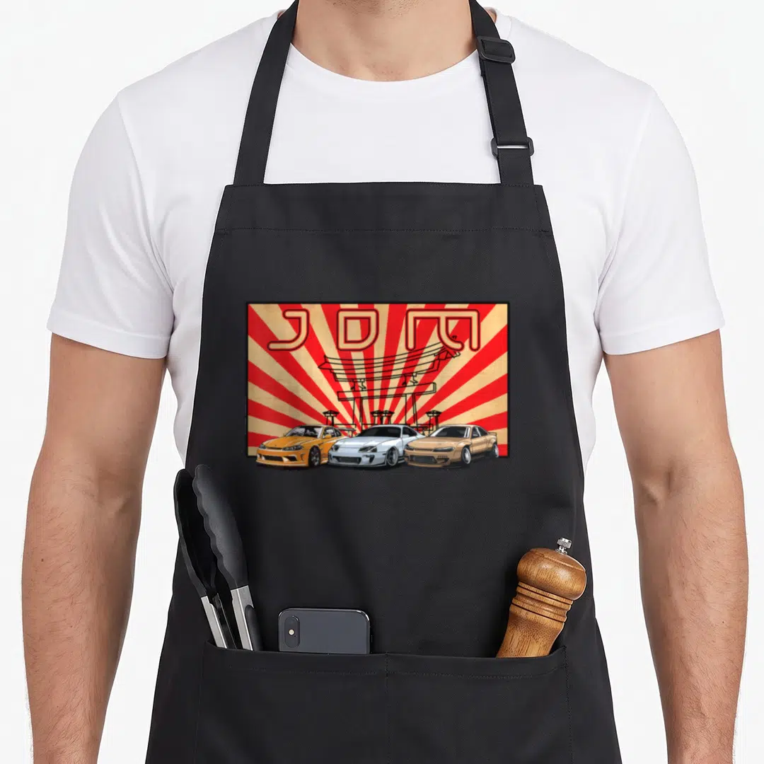 Jdm japanese car apron - Three cartoon cars under a red torii gate with JDM text on a sunburst background — shown on a black cotton canvas apron against a white background