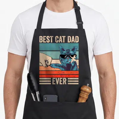 Cat bump father day apron - Blue cat with sunglasses fist bumping a hand over striped background with text BEST CAT DAD — shown on a black cotton canvas apron against a white background
