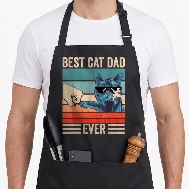 Cat bump father day apron - Blue cat with sunglasses fist bumping a hand over striped background with text BEST CAT DAD — shown on a black cotton canvas apron against a white background