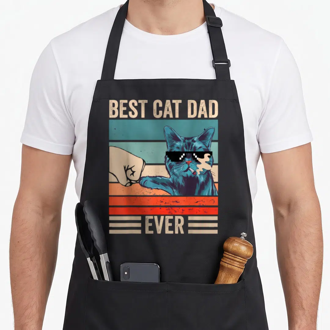 Cat bump father day apron - Blue cat with sunglasses fist bumping a hand over striped background with text BEST CAT DAD — shown on a black cotton canvas apron against a white background