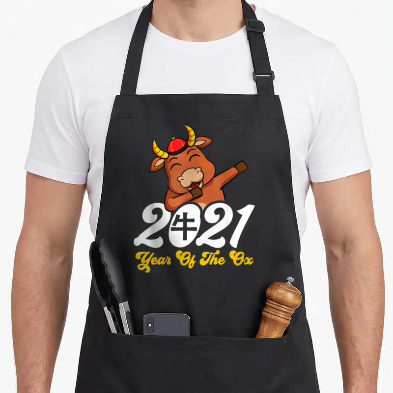 Chinese horoscope zodiac aprons - Cartoon ox in red hat behind white 2021 text and yellow script — shown on a black cotton canvas apron against a white background