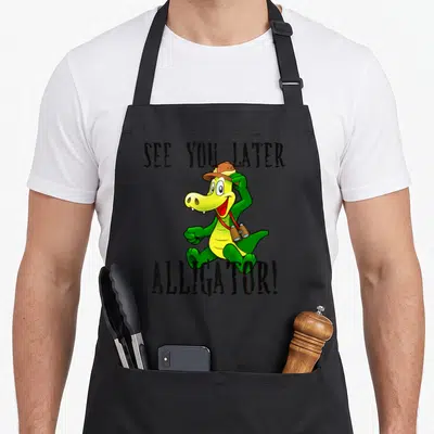 See later apron - Cartoon green alligator with brown hat and binoculars between black text — shown on a black cotton canvas apron against a white background