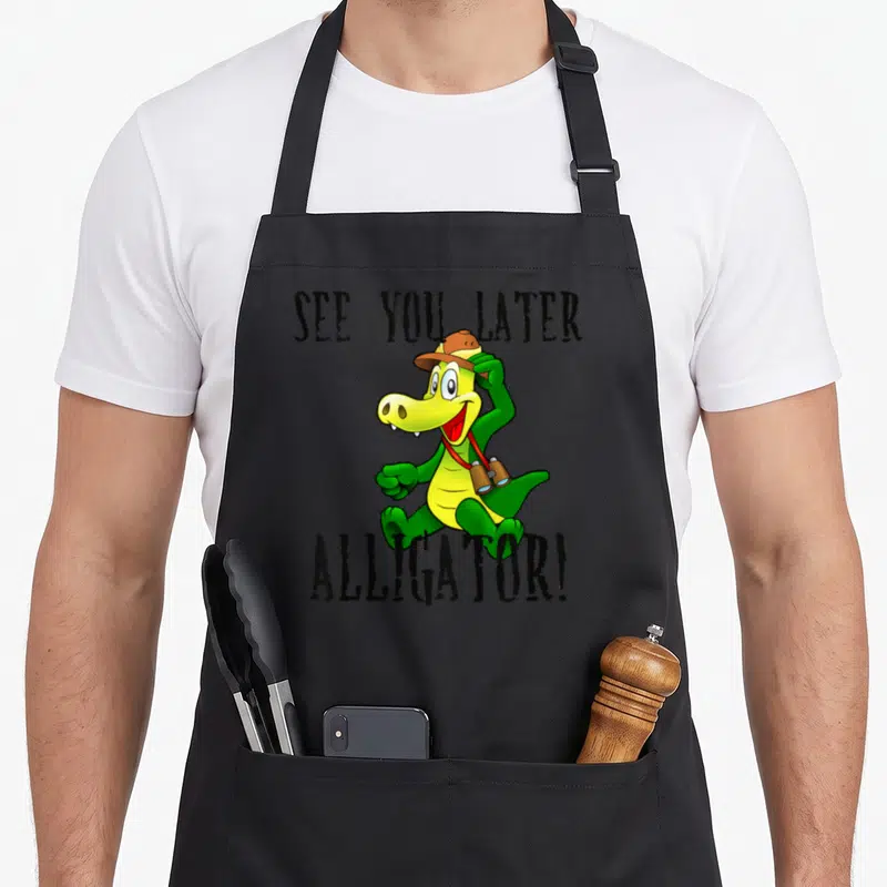 See later apron - Cartoon green alligator with brown hat and binoculars between black text — shown on a black cotton canvas apron against a white background