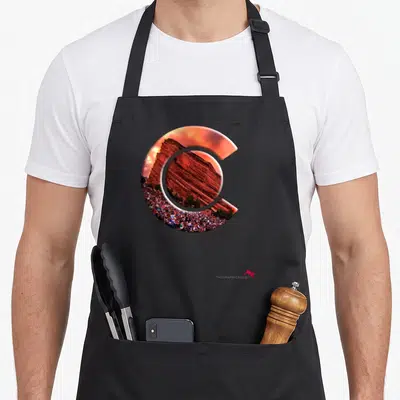 Rock amphitheater colorado apron - Letter C shape filled with red rock formation, crowd, and orange sky photograph — shown on a black cotton canvas apron against a white background