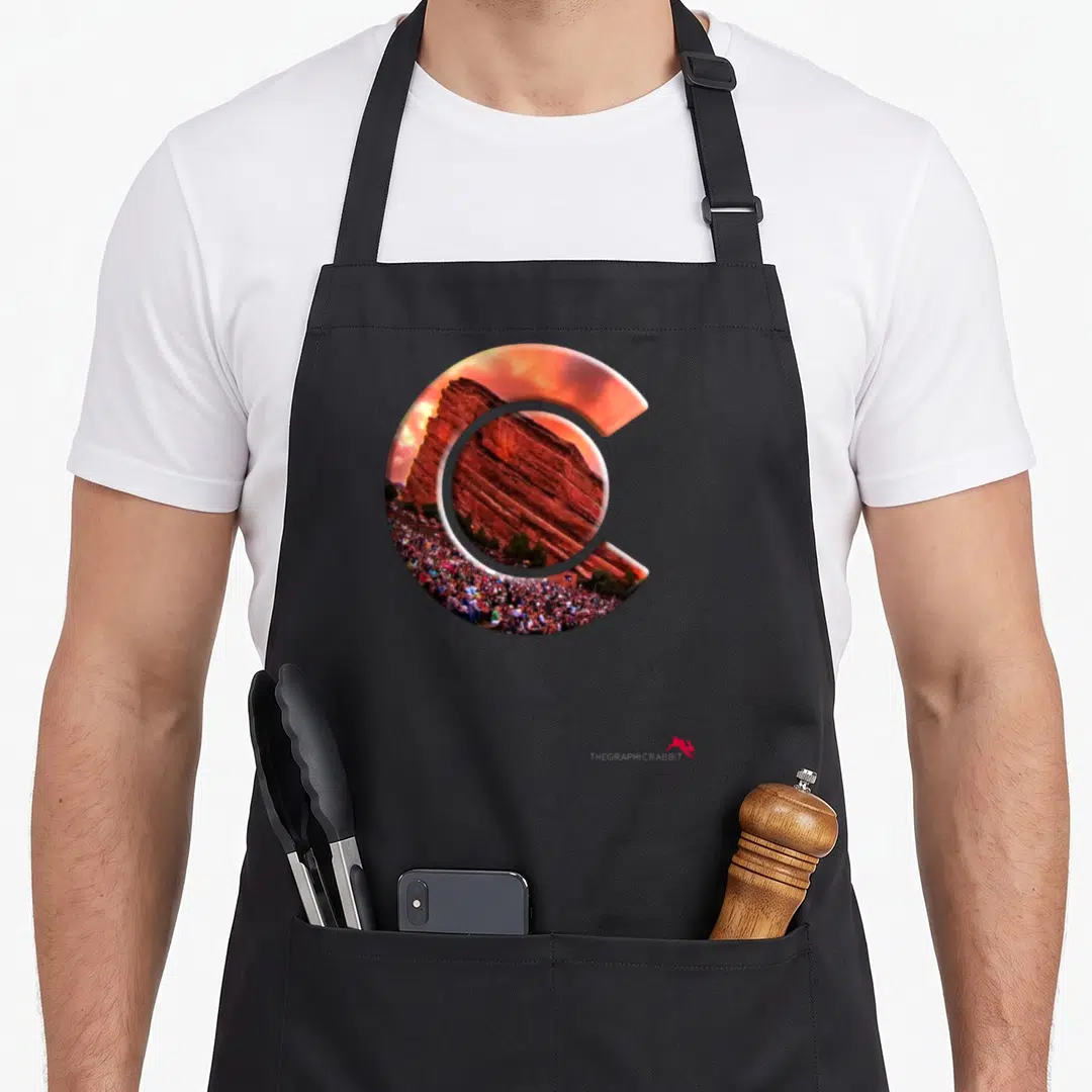 Rock amphitheater colorado apron - Letter C shape filled with red rock formation, crowd, and orange sky photograph — shown on a black cotton canvas apron against a white background