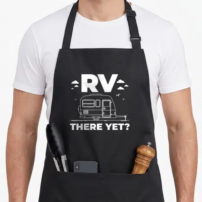 Yet trailer apron - White line-art RV with clouds and text RV THERE YET? on dark background — shown on a black cotton canvas apron against a white background