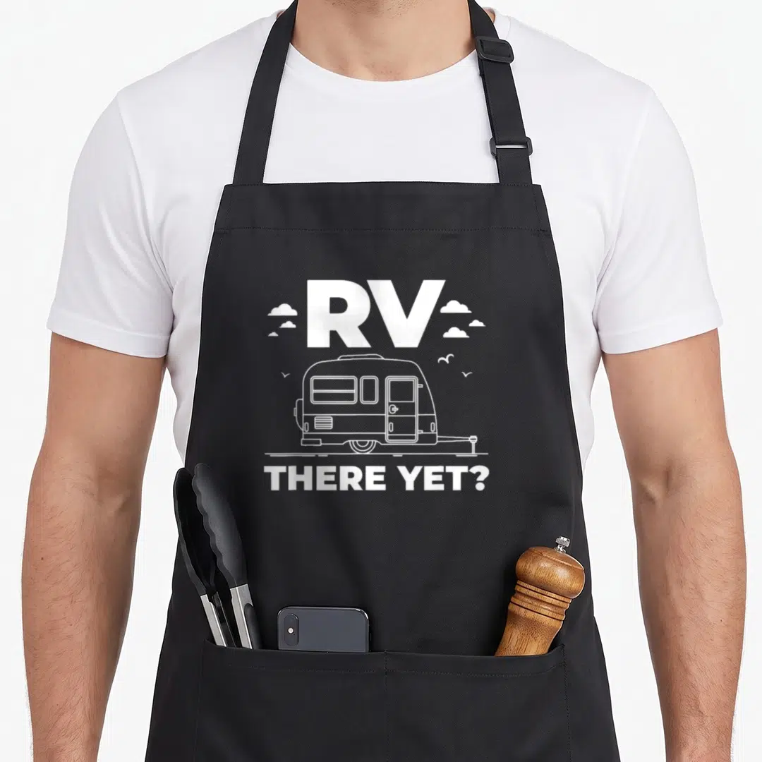 Yet trailer apron - White line-art RV with clouds and text RV THERE YET? on dark background — shown on a black cotton canvas apron against a white background