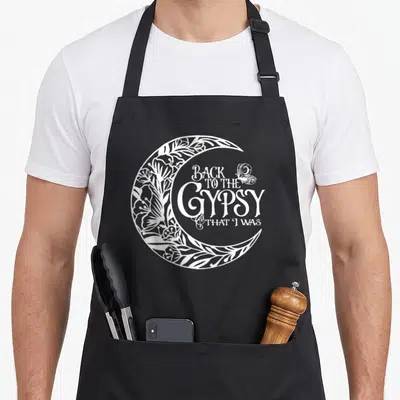 Funny gypsy apron - White crescent moon with floral patterns and text Back to the Gypsy That I Was — shown on a black cotton canvas apron against a white background
