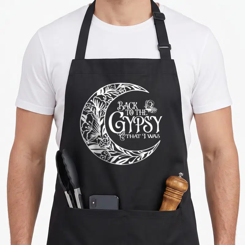 Funny gypsy apron - White crescent moon with floral patterns and text Back to the Gypsy That I Was — shown on a black cotton canvas apron against a white background