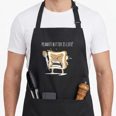 Gag butter apron - Angry cartoon toast holding knife with white splatters and text PEANUT BUTTER IS LIFE — shown on a black cotton canvas apron against a white background