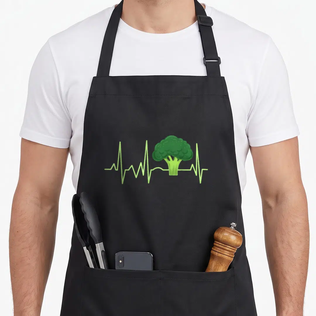 Heartbeat broccoli apron - Green cartoon broccoli floret integrated into a horizontal light green heartbeat pulse line — shown on a black cotton canvas apron against a white background