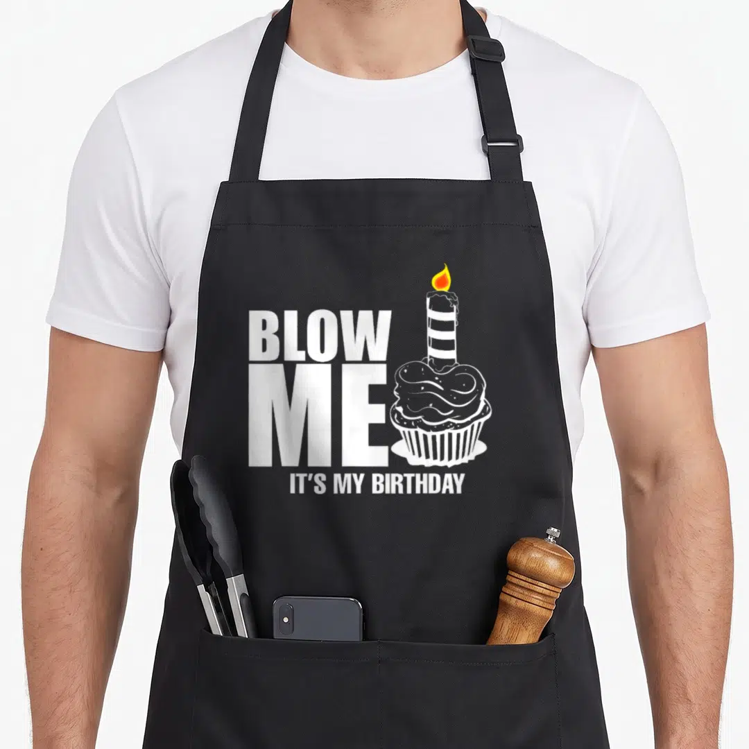 Birthday cake apron - BLOW ME IT'S MY BIRTHDAY text next to a cartoon cupcake with a lit candle — shown on a black cotton canvas apron against a white background