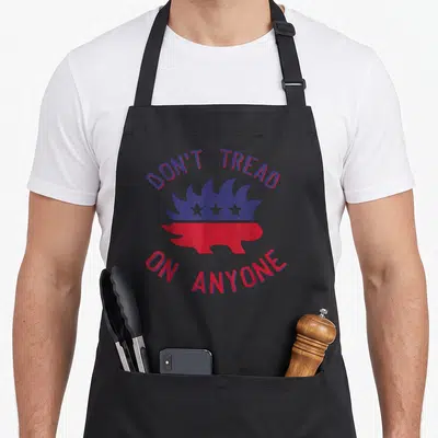 Tread porcupine apron - Minimalist hedgehog split blue and red with DON'T TREAD, ON ANYONE text arching above and below — shown on a black cotton canvas apron against a white background