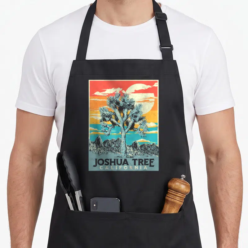 Joshua artistic apron - Retro blue Joshua tree against red orange yellow and blue sunset bands with California text — shown on a black cotton canvas apron against a white background