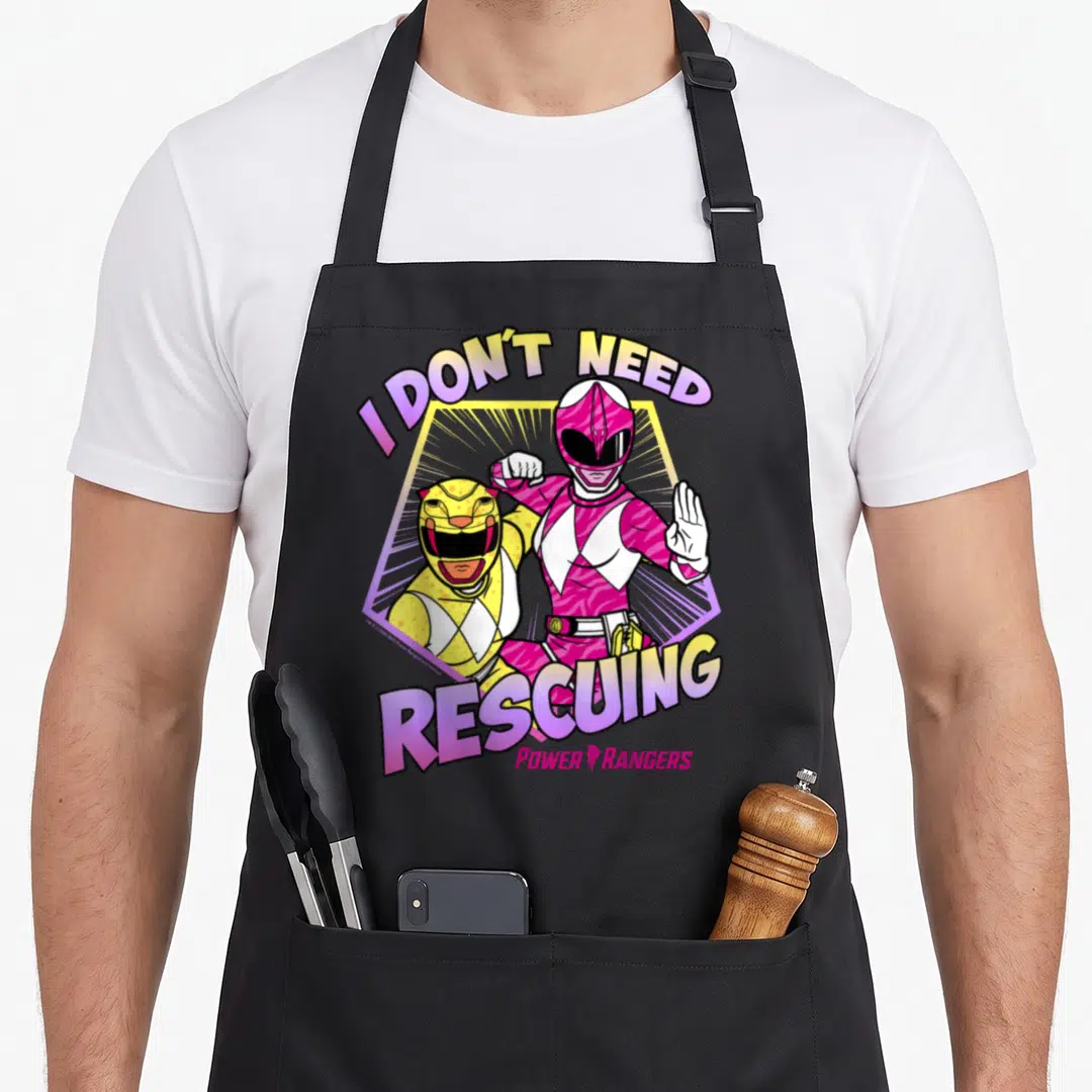 Rescuing rangers apron - Two Power Rangers in pink and yellow suits inside a hexagon with I Don't Need Rescuing text — shown on a black cotton canvas apron against a white background