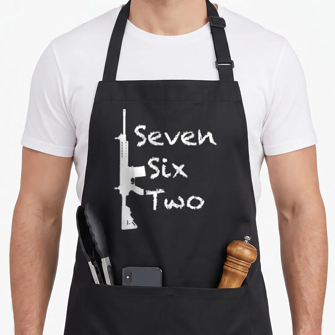 Funny six seven apron - White rifle silhouette beside stacked Seven Six Two text — shown on a black cotton canvas apron against a white background