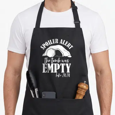 Tomb empty he is risen apron - White open tomb silhouette with Spoiler Alert and Luke 24:24 text — shown on a black cotton canvas apron against a white background