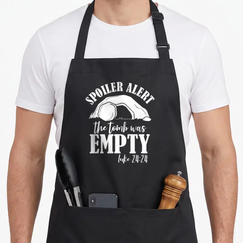 Tomb empty he is risen apron - White open tomb silhouette with Spoiler Alert and Luke 24:24 text — shown on a black cotton canvas apron against a white background
