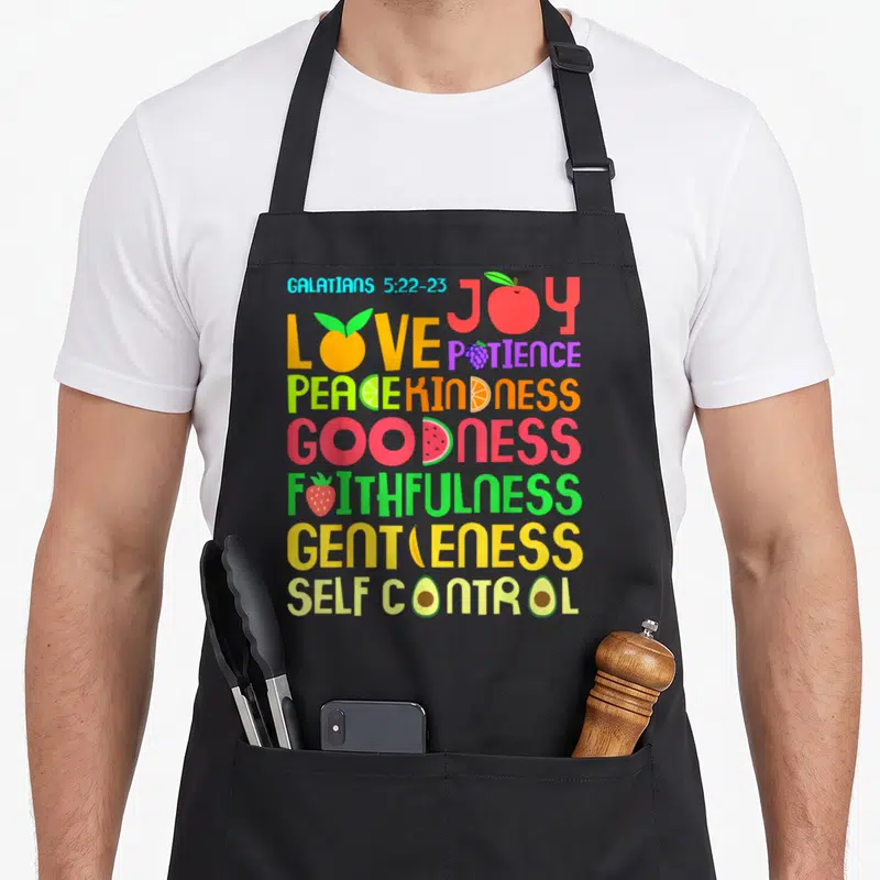 Funny bible verse apron - Stacked colorful text Galatians 5:22-23 with fruit icons replacing letters in Joy Love Peace — shown on a black cotton canvas apron against a white background