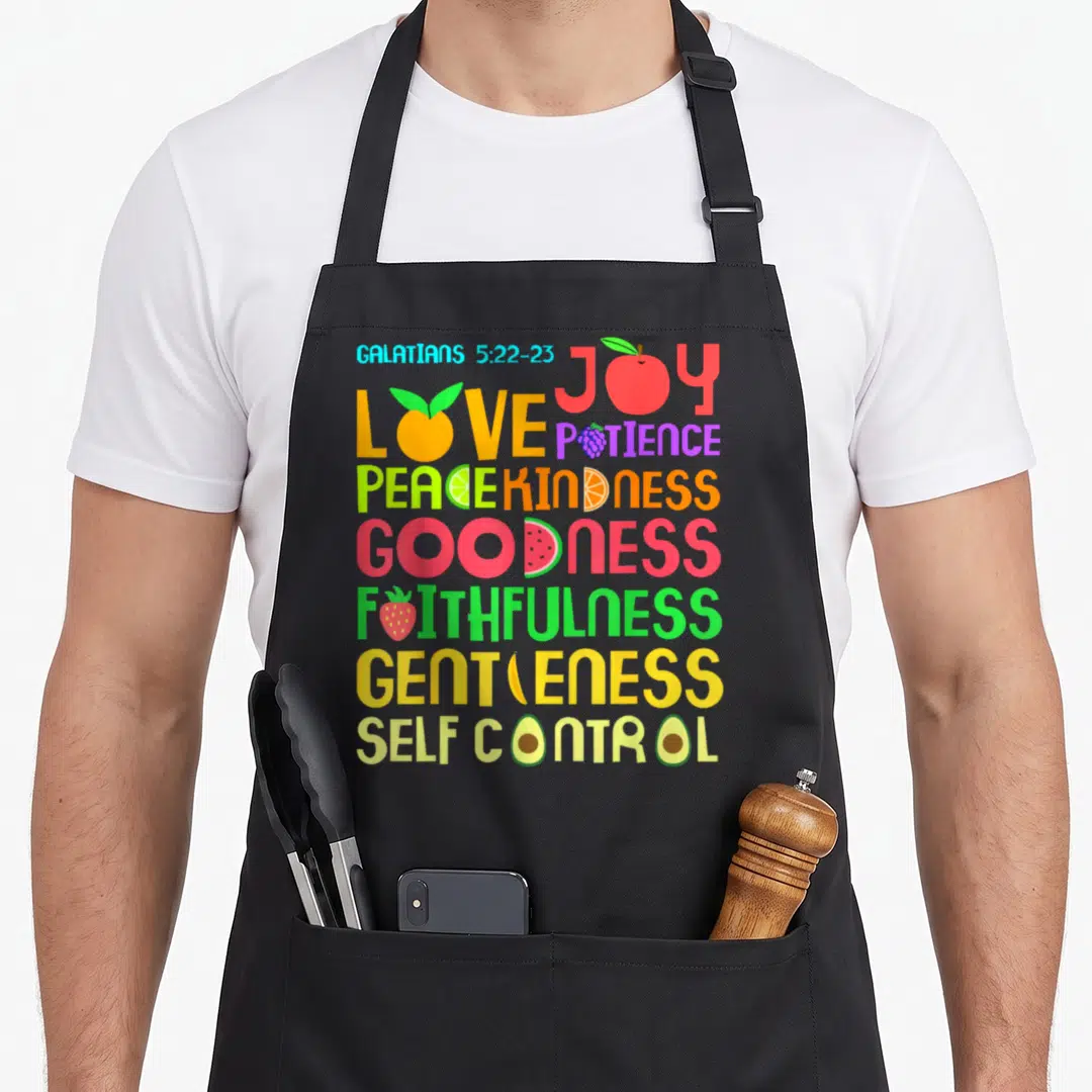 Funny bible verse apron - Stacked colorful text Galatians 5:22-23 with fruit icons replacing letters in Joy Love Peace — shown on a black cotton canvas apron against a white background
