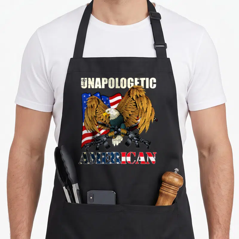 Unapologetic eagle american flag apron - Bald eagle holding two rifles in front of a waving flag with unapologetic — shown on a black cotton canvas apron against a white background