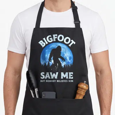 Nobody bigfoot apron - Bigfoot silhouette inside blue moon with white distressed text BIGFOOT SAW ME BUT NOBODY — shown on a black cotton canvas apron against a white background