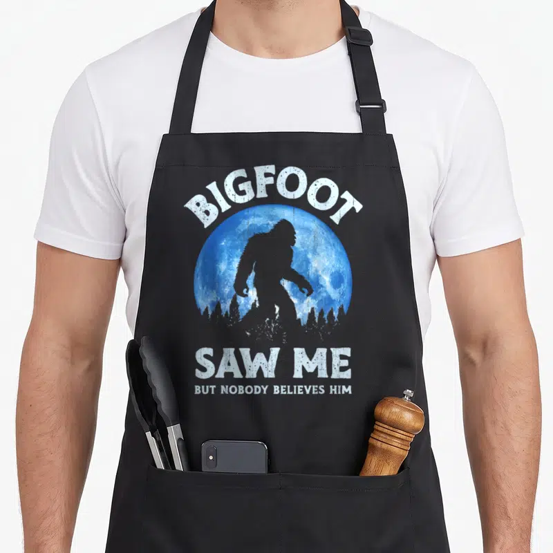 Nobody bigfoot apron - Bigfoot silhouette inside blue moon with white distressed text BIGFOOT SAW ME BUT NOBODY — shown on a black cotton canvas apron against a white background