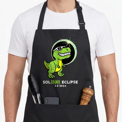 Dino april apron - Green cartoon dinosaur in sunglasses with Sol Rawr Eclipse text and 4 8 2024 date — shown on a black cotton canvas apron against a white background