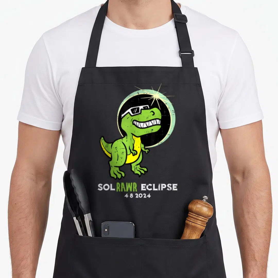 Dino april apron - Green cartoon dinosaur in sunglasses with Sol Rawr Eclipse text and 4 8 2024 date — shown on a black cotton canvas apron against a white background