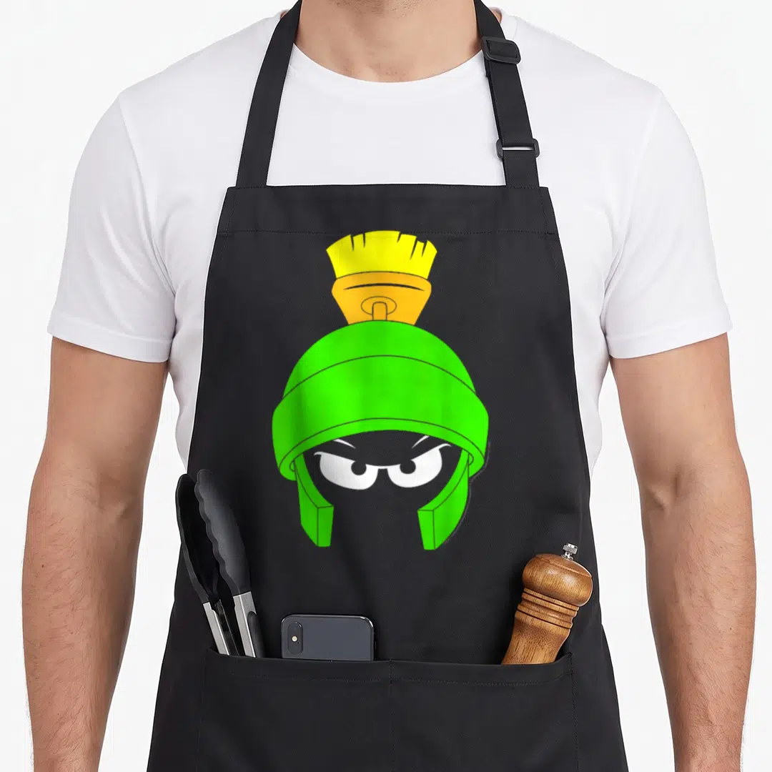 Marvin face apron - Green helmet with yellow orange plume and two white cartoon eyes — shown on a black cotton canvas apron against a white background