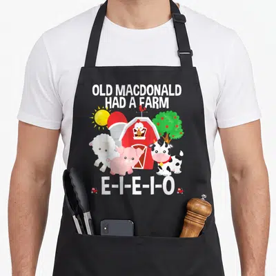 Macdonald farm apron - Cartoon farm scene with sheep, pig, cow, red barn, and OLD MACDONALD HAD A FARM text — shown on a black cotton canvas apron against a white background