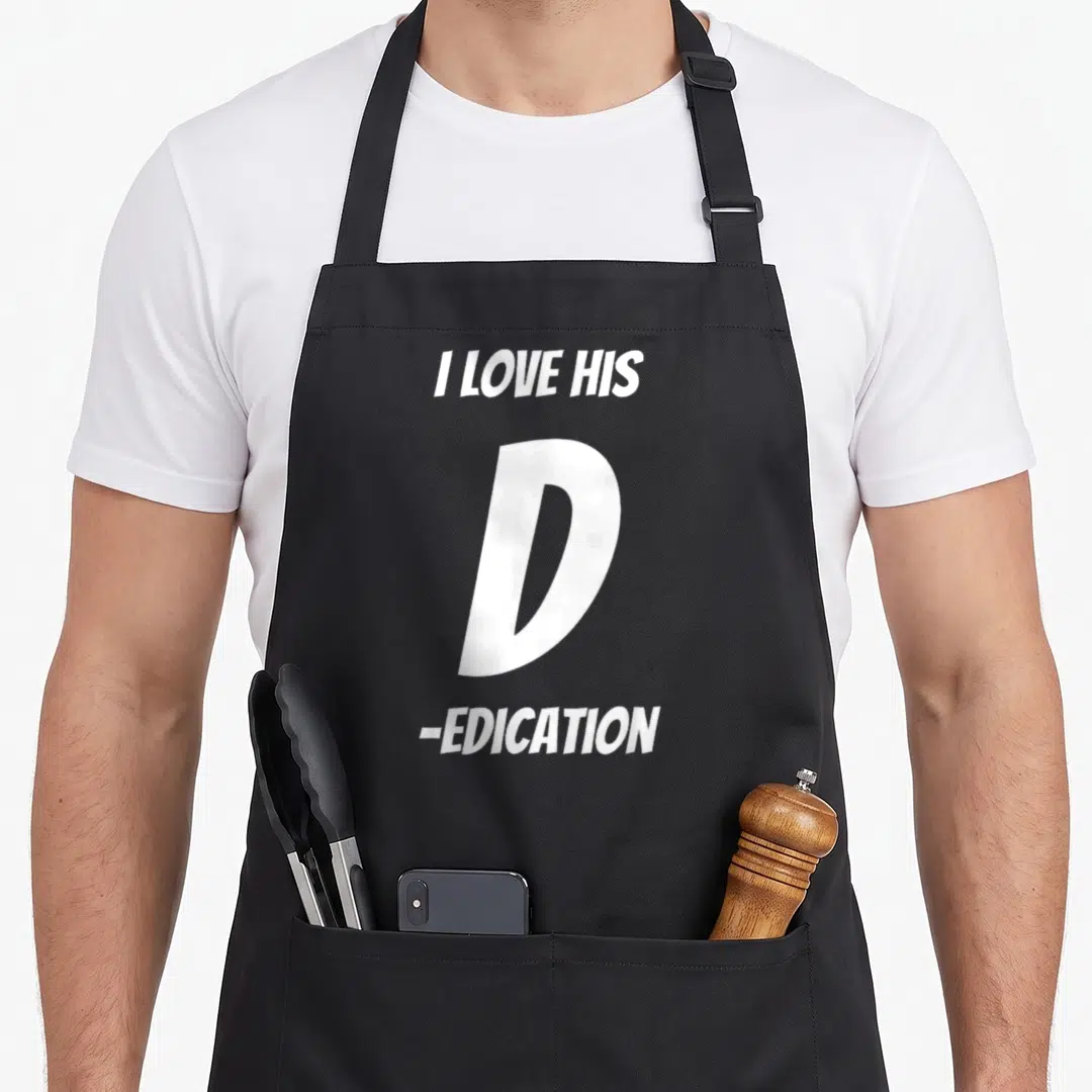 Dedication dirty joke aprons - White stacked text reading I LOVE HIS D EDICATION with large center letter — shown on a black cotton canvas apron against a white background