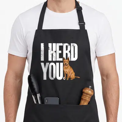 Herd dog kitchen apron - I HERD YOU text with cartoon brown dog sitting beside white letters on black fabric — shown on a black cotton canvas apron against a white background