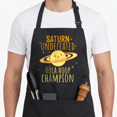 Saturn undefeated apron - Cartoon Saturn planet with rings acting as hula hoops and text reading Hula Hoop Champion — shown on a black cotton canvas apron against a white background