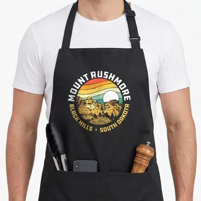 Mount 80s apron - Retro circular Mount Rushmore badge with striped sunset and Black Hills South Dakota text — shown on a black cotton canvas apron against a white background