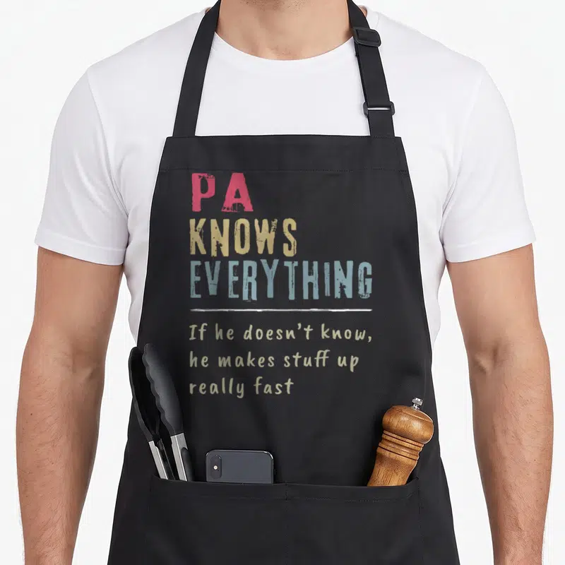 Vintage grandpa apron - Stacked PA KNOWS EVERYTHING text in red yellow blue and tan with white divider line — shown on a black cotton canvas apron against a white background