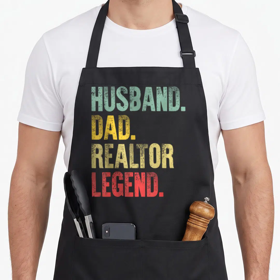 Realtor husband apron - Stacked text HUSBAND DAD REALTOR LEGEND in green yellow red distressed font — shown on a black cotton canvas apron against a white background