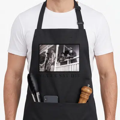 Die novelty apron - Black and white photo of kids on porch with NEVER SAY DIE text below — shown on a black cotton canvas apron against a white background