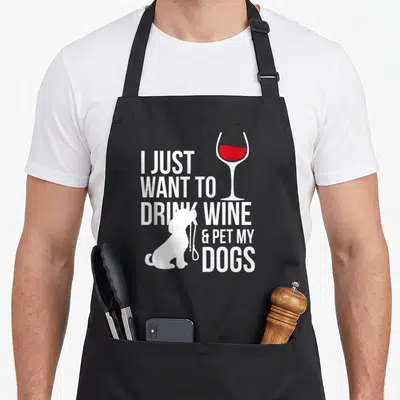 Drink wine dog apron - White stacked text I just want to drink wine and pet my dogs with red wine glass and dog — shown on a black cotton canvas apron against a white background