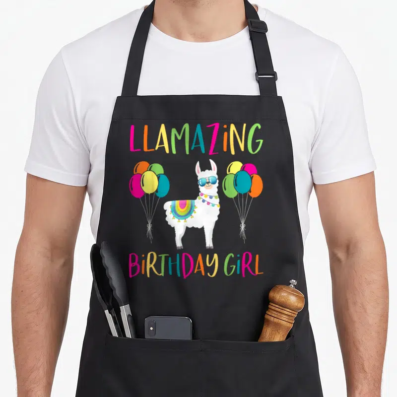 Birthday alpaca apron - Cartoon white llama with sunglasses and rainbow blanket between balloon clusters and LLAMAZING — shown on a black cotton canvas apron against a white background