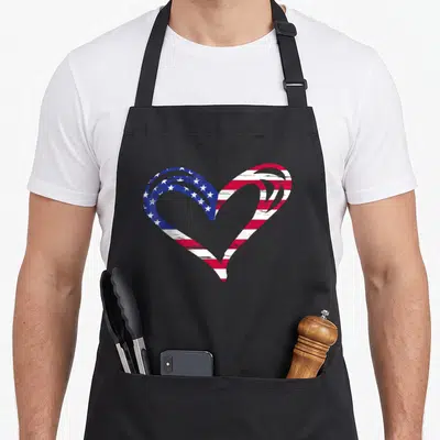 Armed american flag apron - Distressed heart shape filled with blue white and red USA flag pattern — shown on a black cotton canvas apron against a white background