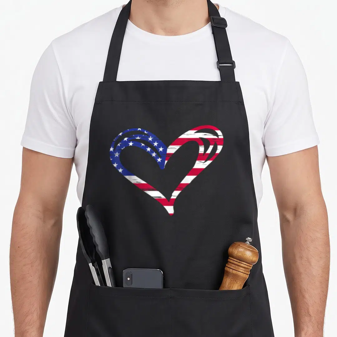 Armed american flag apron - Distressed heart shape filled with blue white and red USA flag pattern — shown on a black cotton canvas apron against a white background