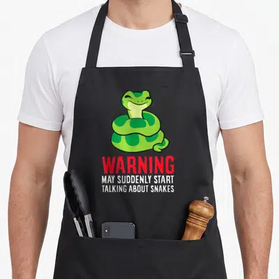 May talk about apron - Cartoon green coiled snake above red warning text and white slogan lines — shown on a black cotton canvas apron against a white background