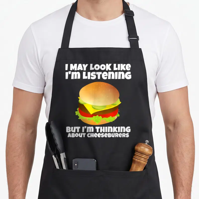 Listening cheeseburgers apron - Cartoon cheeseburger with white text reading I may look like I'm listening but I'm — shown on a black cotton canvas apron against a white background