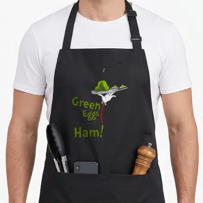 Vintage ham egg apron - Hand holding plate of green ham and eggs with stacked text Green Eggs and Ham — shown on a black cotton canvas apron against a white background